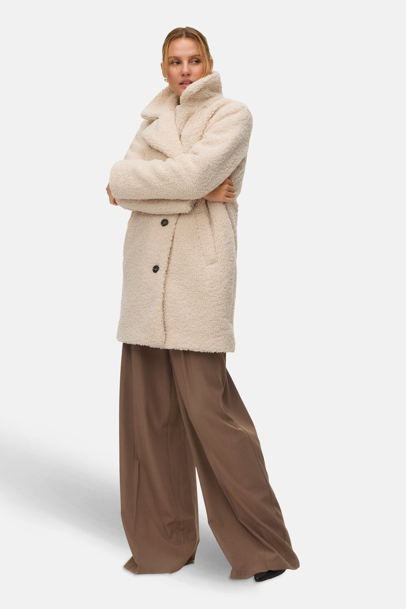 Veste longue beige de VERO MODA avec texture de type peau de mouton, portée avec un pantalon marron ample.
