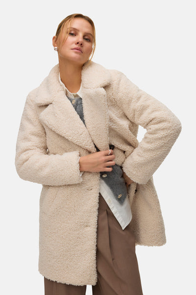 Veste longue beige de VERO MODA avec texture moutonnée, portée sur chemise blanche et pantalon marron.