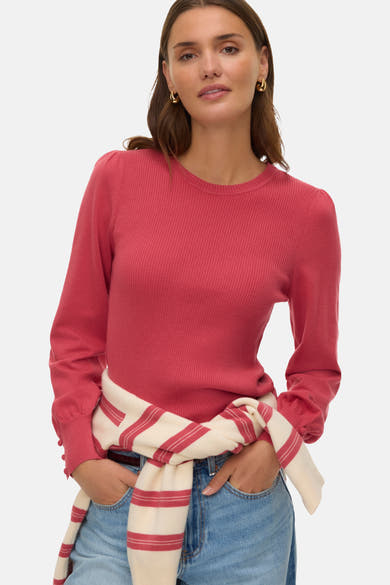Pull à col rond rose de VERO MODA, en maille côtelée, porté avec un pull rayé noué à la taille et un jean bleu.
