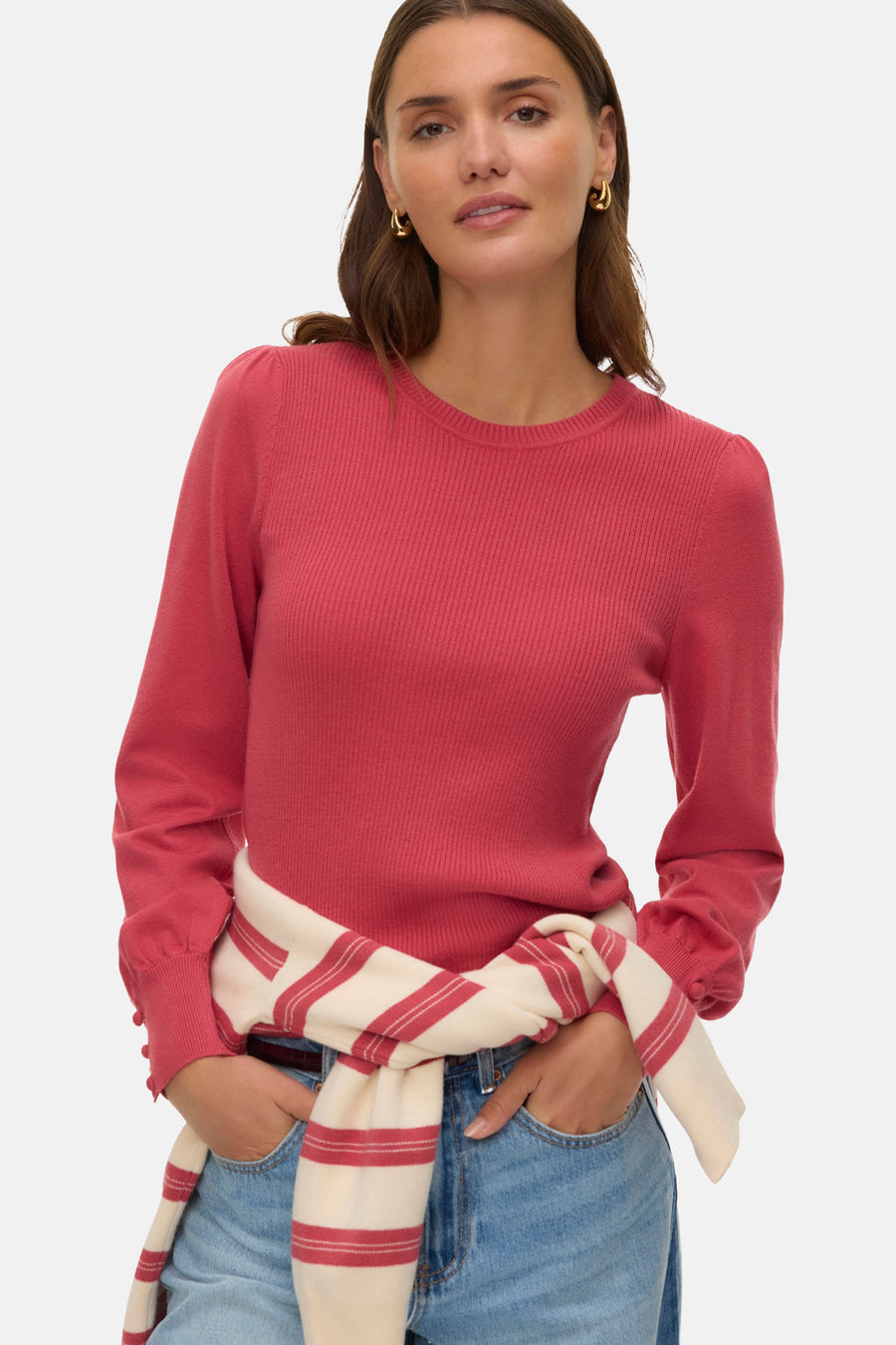 Pull à col rond rose de VERO MODA, en maille côtelée, porté avec un pull rayé noué à la taille et un jean bleu.
