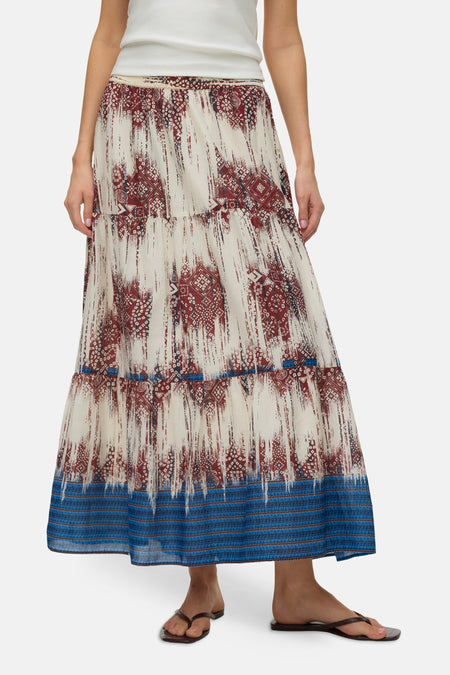 Jupe longue multicolore de VERO MODA® à motif abstrait rouge rouille et bleu sur fond crème, portée avec un débardeur blanc et des sandales.