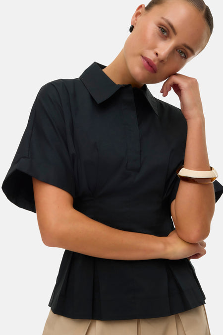 Blouse noire de VERO MODA avec col polo, patte de boutonnage courte et manches amples, portée par une femme avec un bracelet bicolore.