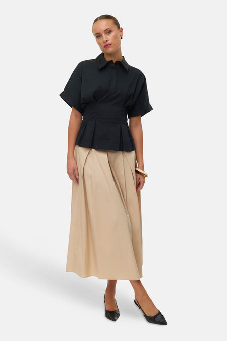 Blouse à manches courtes noire de VERO MODA avec ourlet plissé, portée avec une jupe longue beige et des chaussures noires.