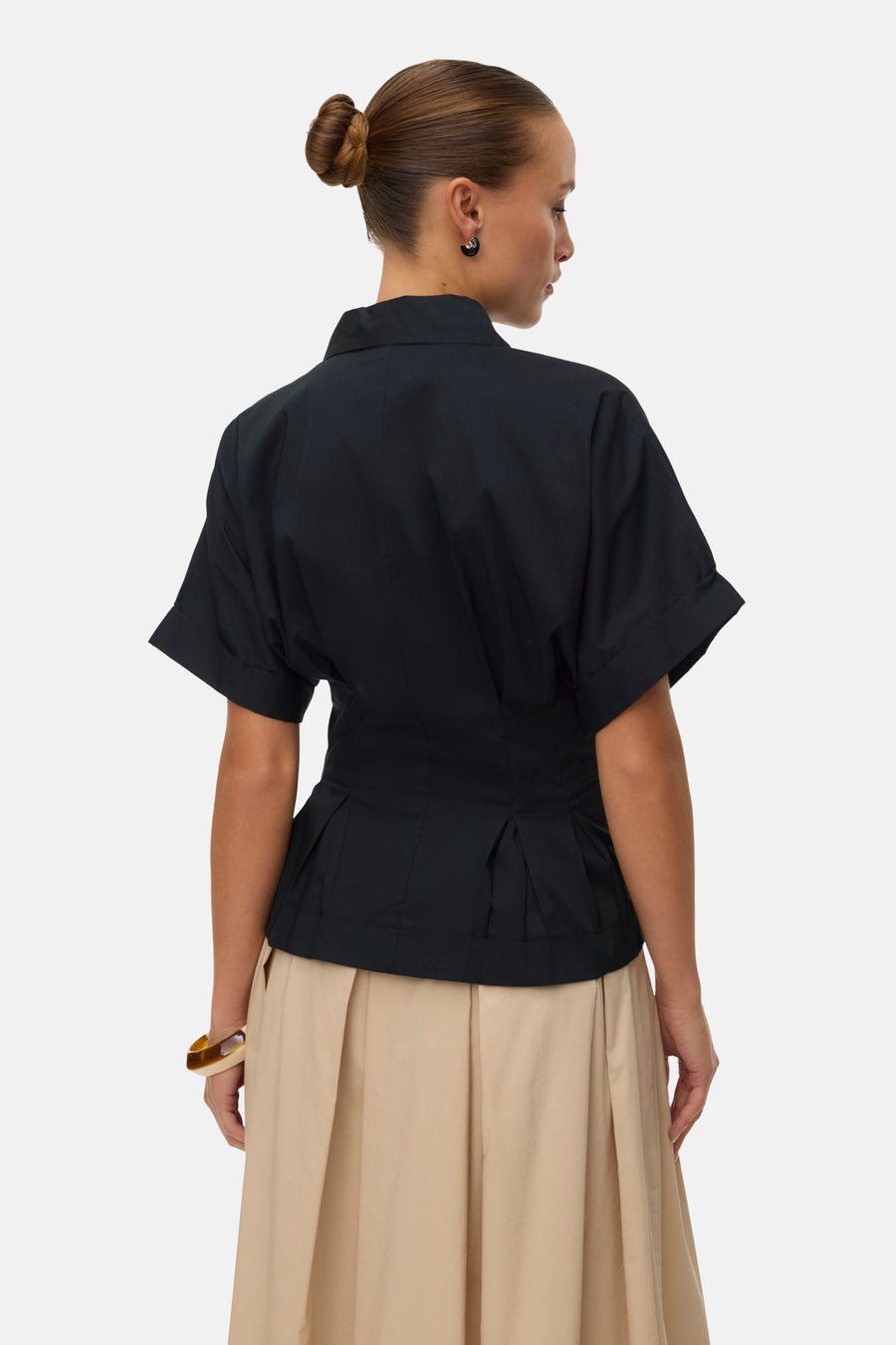Blouse noire à manches courtes de VERO MODA, vue de dos, avec col chemise, taille cintrée et plis, portée avec une jupe plissée beige et un bracelet.