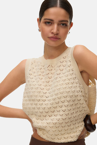 Débardeur écru de VERO MODA® en maille ajourée, porté avec un pantalon marron et des boucles d'oreilles argentées.