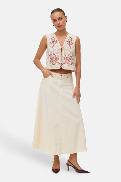 Gilet écru brodé de VERO MODA avec motifs feuilles roses, liens noués, porté sur une jupe longue écrue et escarpins bordeaux.