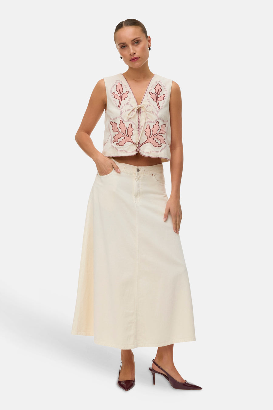 Gilet écru brodé de VERO MODA avec motifs feuilles roses, liens noués, porté sur une jupe longue écrue et escarpins bordeaux.