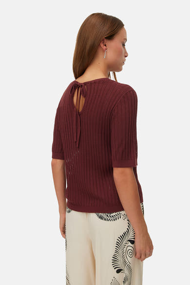 Pull col rond brun en maille ajourée de VERO MODA, avec nœud décoratif au dos, porté sur un pantalon à motifs.
