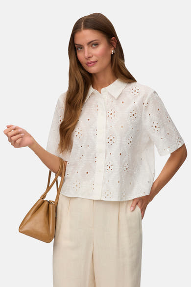Chemise à manches courtes blanche de VERO MODA® en broderie anglaise à motifs floraux ajourés, portée avec un pantalon beige et un sac marron.