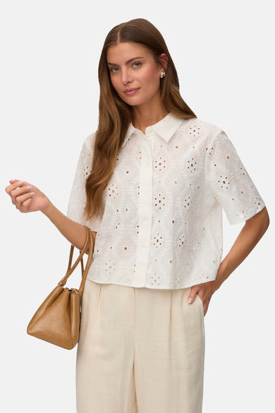 Chemise à manches courtes blanche de VERO MODA® en broderie anglaise à motifs floraux ajourés, portée avec un pantalon beige et un sac marron.