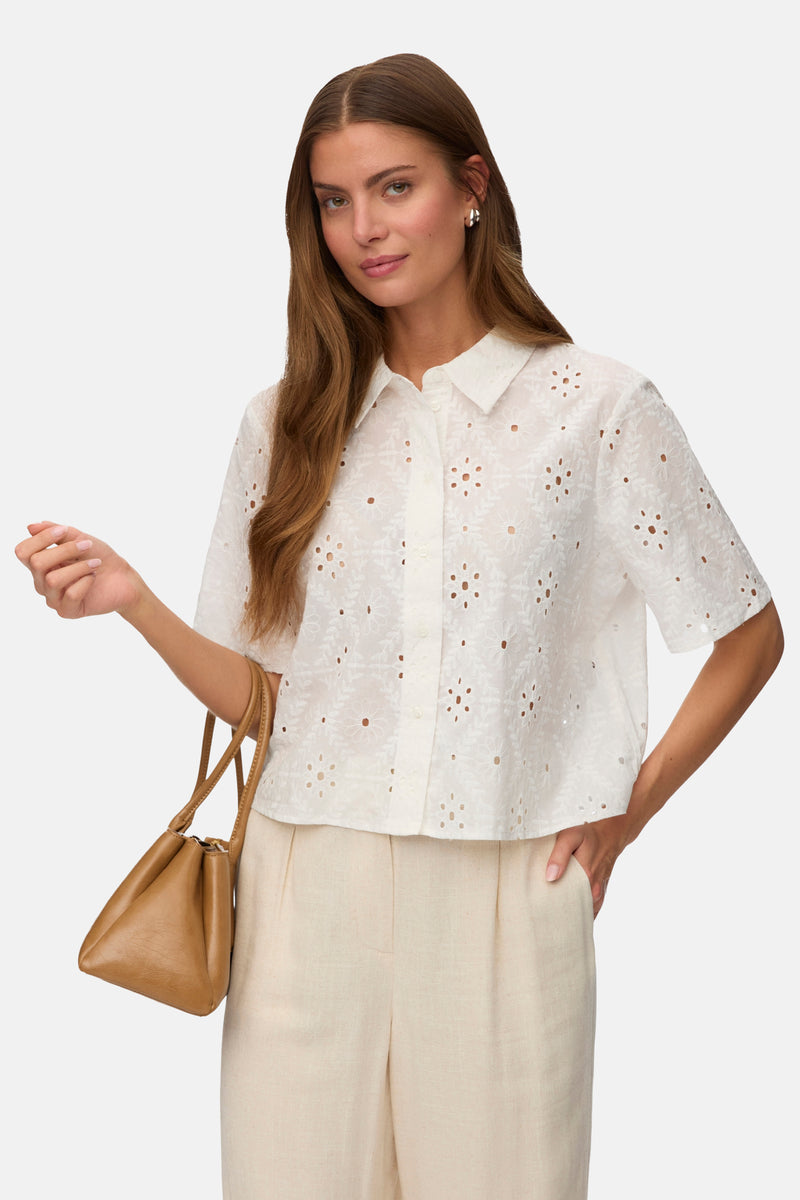 Chemise à manches courtes blanche de VERO MODA® en broderie anglaise à motifs floraux ajourés, portée avec un pantalon beige et un sac marron.