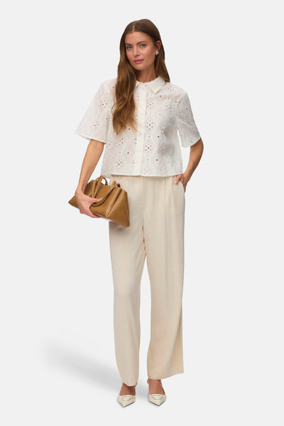 Chemise à manches courtes blanche de VERO MODA, avec broderie anglaise, portée avec un pantalon large beige et des escarpins.