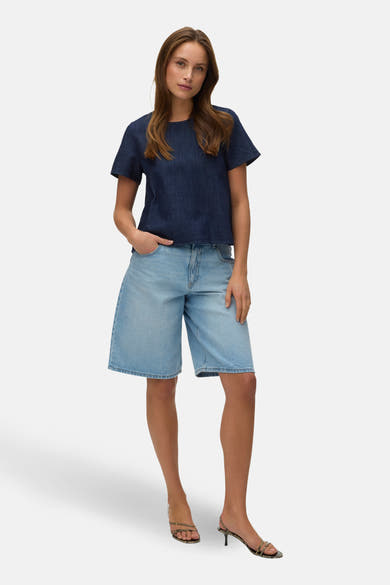 Blouse à manches courtes en denim bleu foncé de VERO MODA, portée avec un short en jean et des sandales à talons.