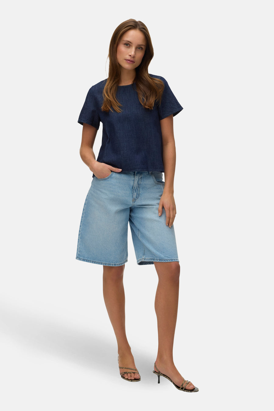 Blouse à manches courtes en denim bleu foncé de VERO MODA, portée avec un short en jean et des sandales à talons.