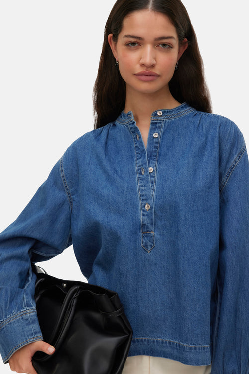 Medium blue denim long-sleeve blouse with button placket and relaxed fit, worn with black bag, front view