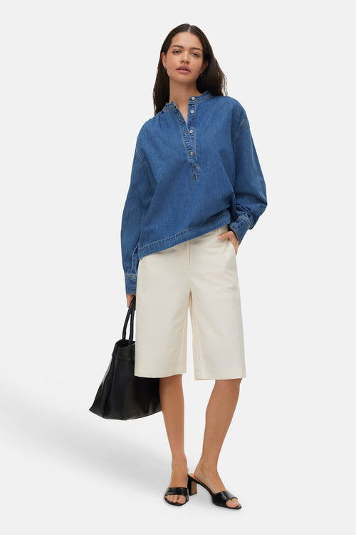 Medium blue denim long-sleeve blouse with button placket, worn with cream bermuda shorts and black mules, full-length lifestyle view