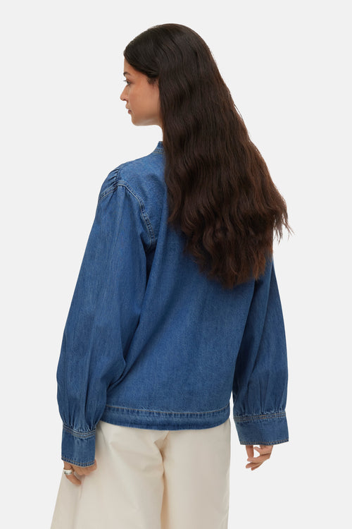 Medium blue denim long-sleeve blouse with gathered shoulders and cuffed sleeves, shown from back view on model with wavy dark hair and cream pants.