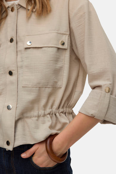 Beige gilet van VERO MODA® met gouden knopen, borstzakken, een verstelbare taille en opgerolde mouwen, gedragen over een spijkerbroek en een houten armband.