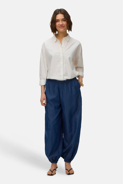 Pantalon bleu de VERO MODA, avec taille élastique et jambes bouffantes, porté avec une chemise blanche boutonnée et des sandales.