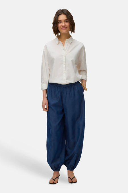 Pantalon bleu de VERO MODA, avec taille élastique et jambes bouffantes, porté avec une chemise blanche boutonnée et des sandales.