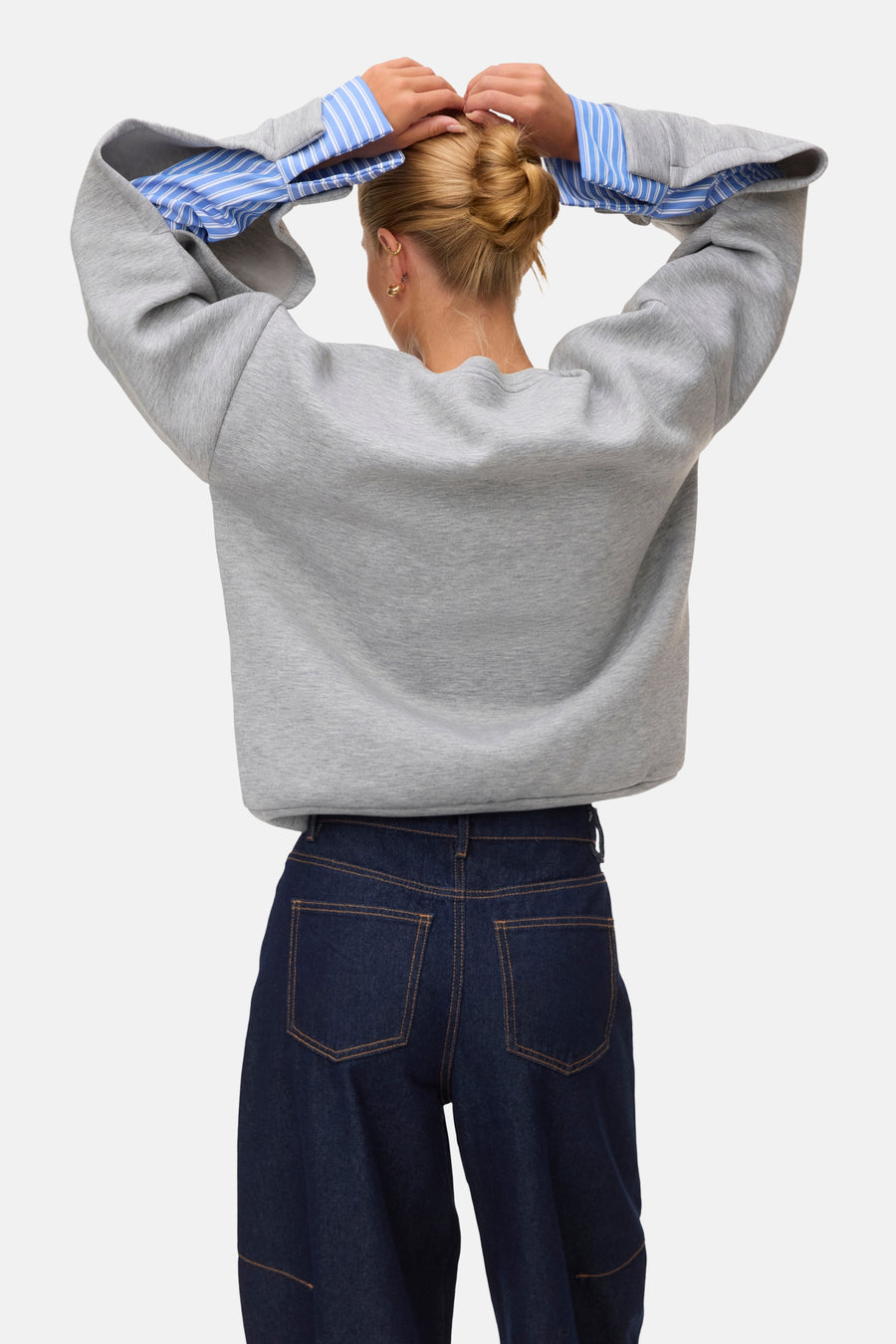 Light grey melange sweatshirt with ribbed cuffs and relaxed fit, shown from back on model wearing blue striped shirt sleeves and dark denim pants