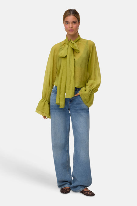 Blouse verte à manches longues avec col lavallière et poignets volantés, portée avec un jean.