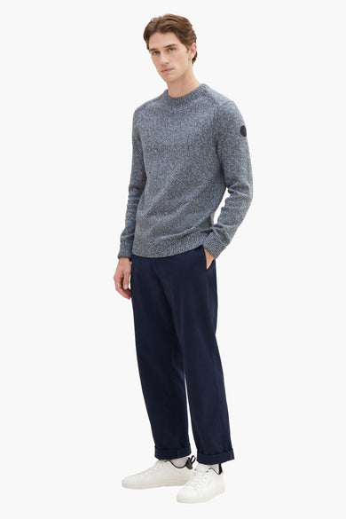 Grey marled crewneck sweater with ribbed cuffs and hem, featuring a small patch on the sleeve, worn with navy trousers and white sneakers in a full-length front view.