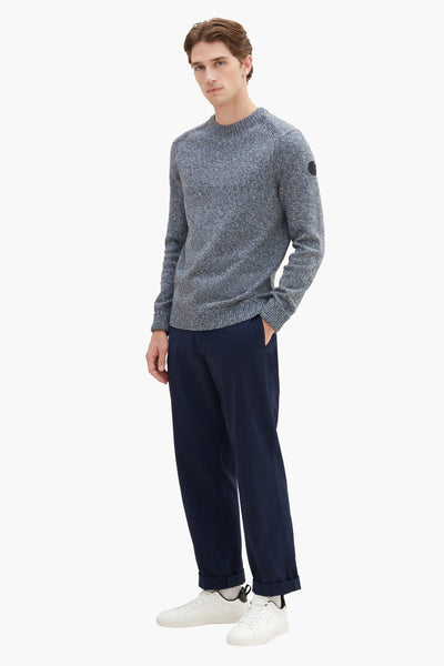 Grey marled crewneck sweater with ribbed cuffs and hem, featuring a small patch on the sleeve, worn with navy trousers and white sneakers in a full-length front view.
