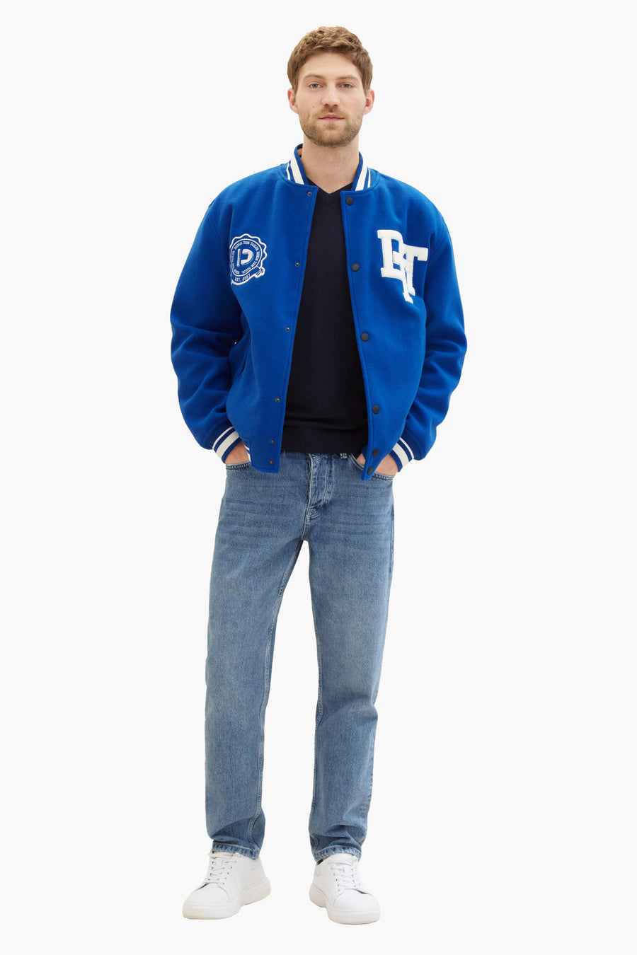 Pull bleu à col V de Tom Tailor, porté avec une veste universitaire bleue, un jean et des baskets blanches.