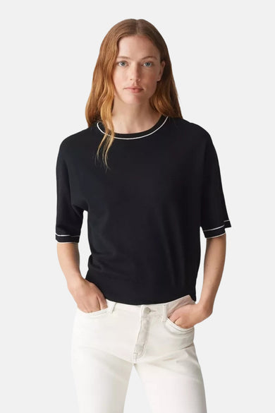 Pull col rond noir de OPUS, avec bordures blanches au col et aux manches, porté avec un jean blanc.
