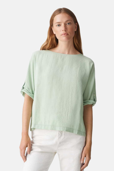 Blouse à manches longues verte de OPUS, avec les manches retroussées et portée avec un pantalon blanc.
