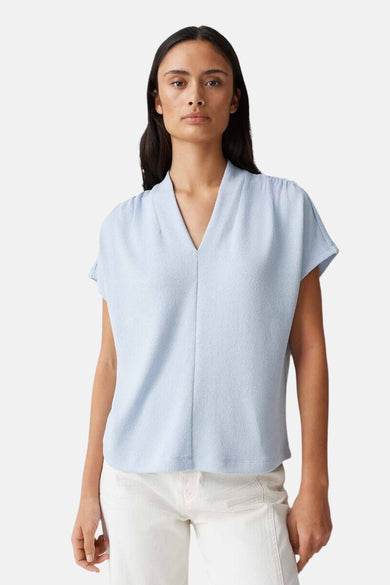Blauwe blouse met korte mouwen van OPUS, met V-hals en licht gekreukte textuur, gedragen met een witte broek.