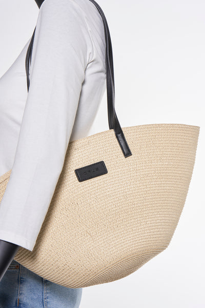 Sac à bandoulière beige en paille de OPUS, avec des anses noires, porté avec un t-shirt blanc et un jean bleu.
