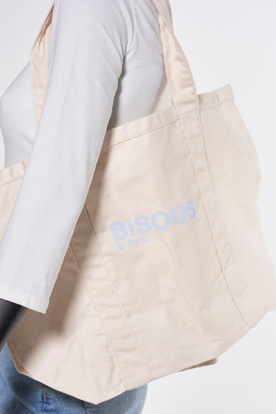 Sac à bandoulière beige de OPUS avec inscription bleu clair, porté sur l'épaule avec un pull blanc et un jean.
