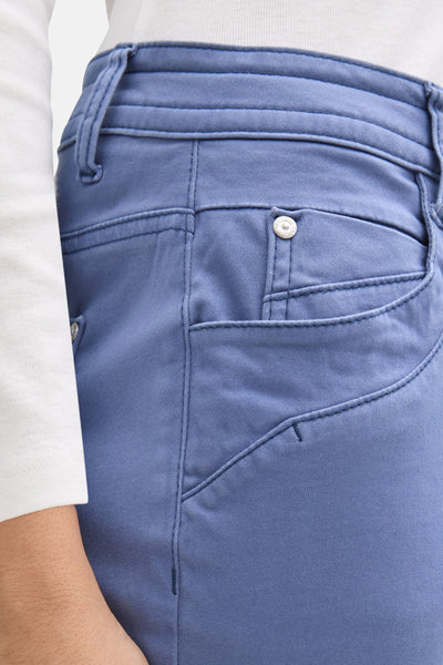 Pantalon coloré bleu de Tom Tailor, avec une poche latérale et une fermeture à bouton, porté avec un haut blanc.

