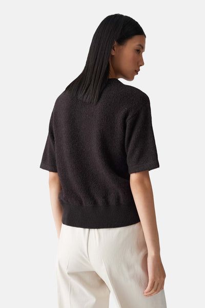 Pull brun par OPUS, à manches courtes et bas côtelé, porté avec un pantalon blanc.