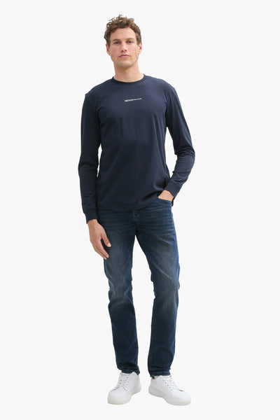 Rechte donkerblauwe jeans van Tom Tailor, gedragen met een marineblauw T-shirt met lange mouwen en witte schoenen.