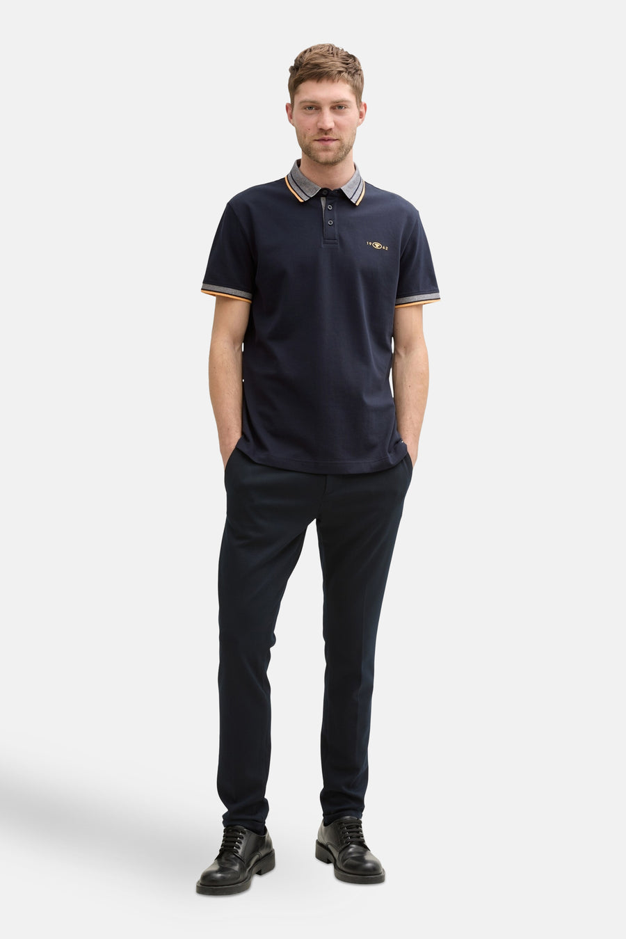 Polo bleu foncé à manches courtes de Tom Tailor, avec rayures sur le col et les manches, porté avec un pantalon bleu marine et des chaussures noires.
