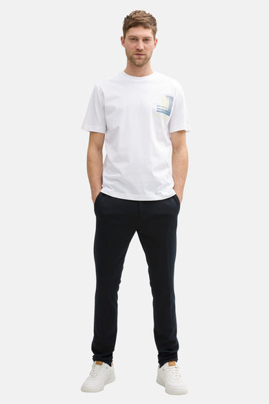 Witte korte mouw T-shirt van Tom Tailor, met een grafisch patroon op de borst. Zwarte broek en witte sneakers.