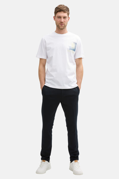 Witte korte mouw T-shirt van Tom Tailor, met een grafisch patroon op de borst. Zwarte broek en witte sneakers.