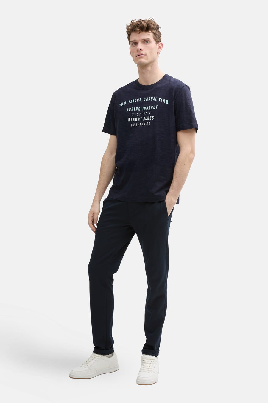 Donkerblauwe T-shirt met korte mouwen van Tom Tailor, met een tekstprint op de voorkant, gedragen met een marineblauwe broek en witte sneakers.