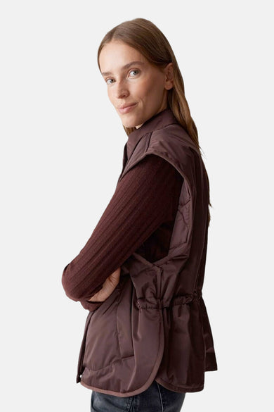 Veste sans manches rouge, par OPUS, avec cordon de serrage à la taille et porté sur un pull marron.
