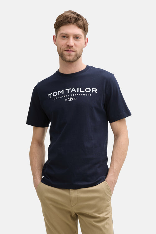 T-shirt bleu à manches courtes de Tom Tailor, avec un imprimé graphique sur le devant, porté avec un pantalon beige.
