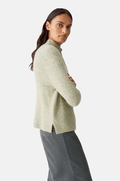 Pull vert d'OPUS, avec une texture douce, porté avec une jupe en velours côtelé gris et un ourlet fendu.