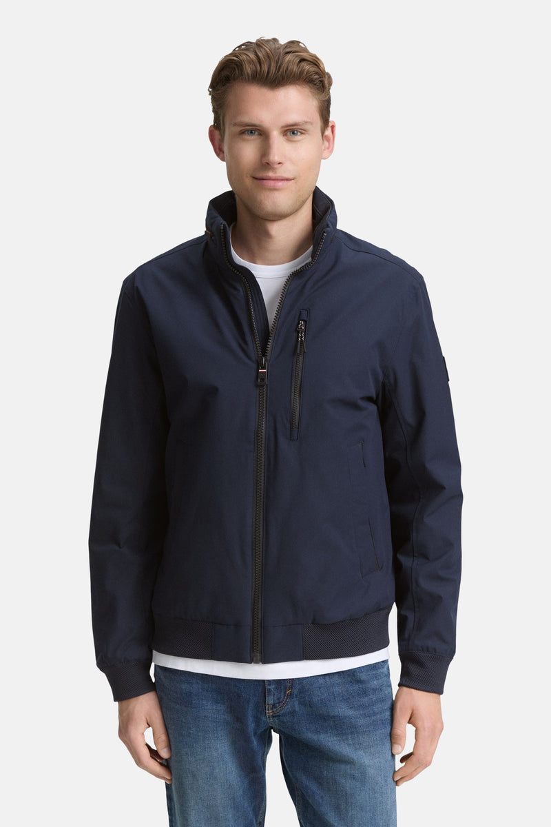 Veste courte bleue par Tom Tailor, avec fermeture éclair visible, portée avec un t-shirt blanc et un jean décontracté.