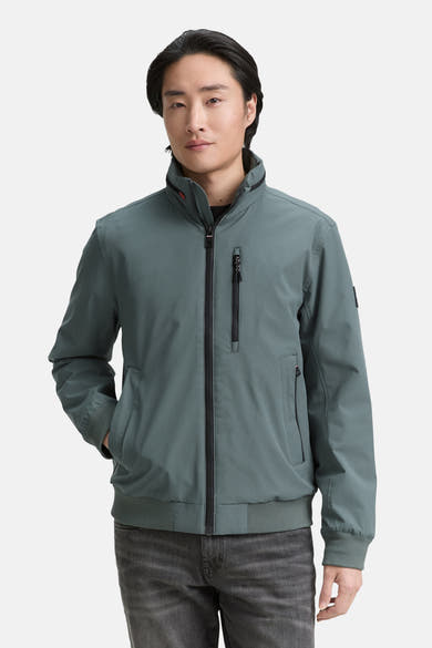 Veste courte verte de Tom Tailor, avec fermeture éclair noire et poches zippées, portée avec un jean gris.
