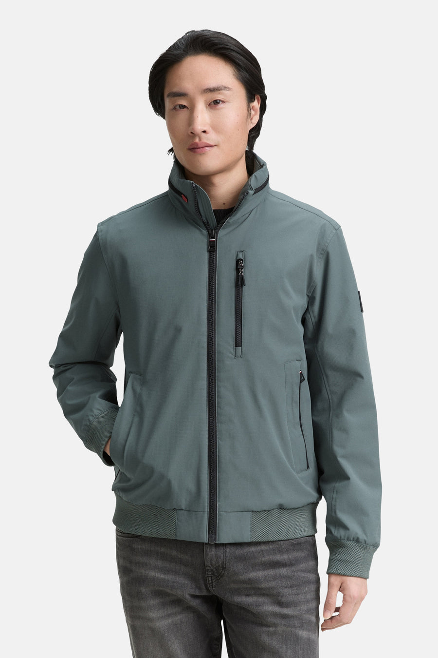 Veste courte verte de Tom Tailor, avec fermeture éclair noire et poches zippées, portée avec un jean gris.
