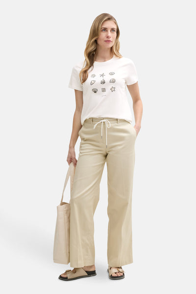 T-shirt à manches courtes blanc de Tom Tailor, avec motifs de coquillages imprimés, porté avec un pantalon beige et un sac fourre-tout.
