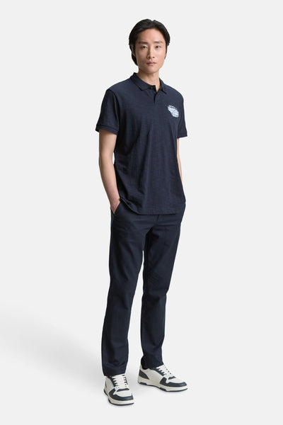 Polo bleu foncé à manches courtes de Tom Tailor, avec un écusson sur la poitrine, porté avec un pantalon et des baskets.