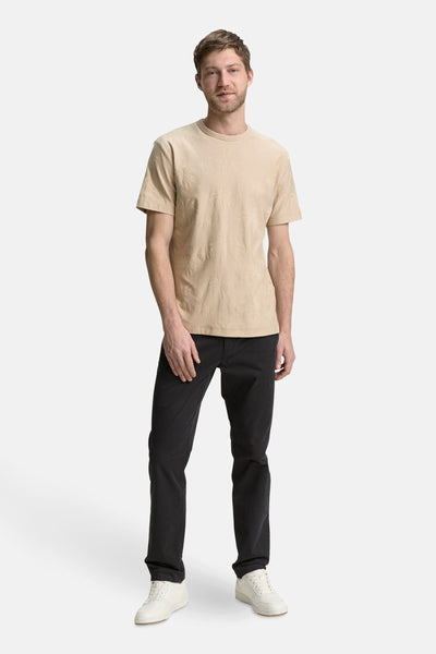 T-shirt à manches courtes beige de Tom Tailor, avec motif de feuilles de palmiers ton sur ton, porté avec un pantalon noir et des baskets blanches.